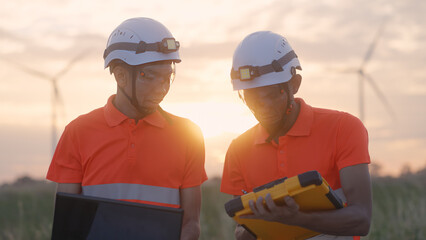 At a wind farm during sunset, two Asian inspectors reviewing data on a laptop and clipboard to analyze system performance and blade rotation. Engineer Wind Turbine.