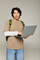 Obraz premium Young fashionable Asian man holds a laptop while standing confidently in a modern urban setting