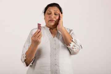 Indian senior woman suffering headache pain while holding pills on her hand isolated on grey studio...