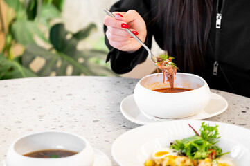 A hand with red nails holds a spoon of soup with herbs, above a white bowl on a table with salad and eggs. Green plants in the background complete this fresh dining setup.