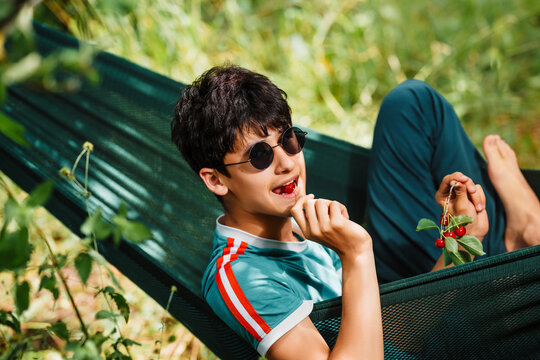 A teenage boy enjoys eat cherries in a hammock in the summer garden.