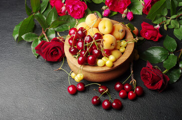 Red cherries and peaches in a wooden bowl on a table surrounded by red roses.