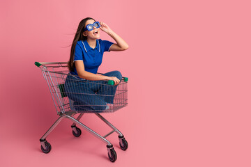 Young cheerful woman sitting in a shopping cart against a pink background wearing casual attire and...