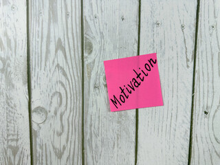 Pink sticky note with Motivation written on it, placed on a white wooden board. This image...