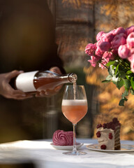 Waiter pours rose wine into glass. Bottle of pink wine with white label. Cake, flowers, wine on festive table. Wine tasting. Alcoholic beverage. Winemaking. Outdoor restaurant. Drink concept.