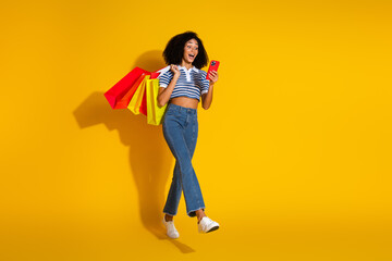 Young fashionable woman carrying shopping bags while using her smartphone against a vibrant yellow background