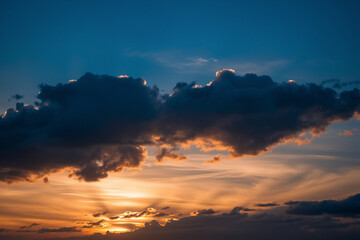 Obraz premium Dramatic dark clouds illuminated by orange sunlight during sunset. The sky blends blue and orange colors, creating a peaceful, scenic, and atmospheric view.