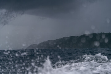A storm approaches Misool Island in Raja Ampat, with dark clouds over the sea. Waves crash, and raindrops blur the view of the misty jungle-covered coastline, creating a dramatic, moody scene.