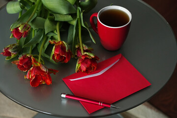Red paper envelope, pen, cup tea and bouquet of red tulips on table close-up