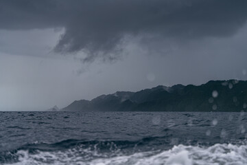 A storm approaches Misool Island in Raja Ampat, with dark clouds over the sea. Waves crash, and raindrops blur the view of the misty jungle-covered coastline, creating a dramatic, moody scene.