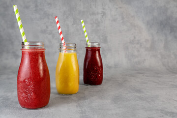 Three glasses of refreshing smoothie with different flavors on a light gray background. Smoothie with strawberries, mango, currants in a glass container with a straw. Concept of trendy cold drinks