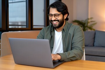 Fototapeta premium Cheerful Bearded Man in Glasses Using Laptop at Wooden Table in Sunlit Contemporary Room