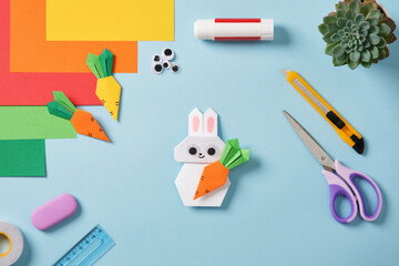 White handmade origami paper rabbit holding a carrot, as well as other paper carrots, tools and supplies for creativity: scissors, cutter, glue, ruler, eraser.