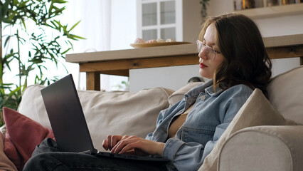 Seated on a sofa, a woman in casual attire with glasses writes in a notebook, her gaze fixed on a laptop screen. This photo emphasizes the dedication and focus required for successful remote work or