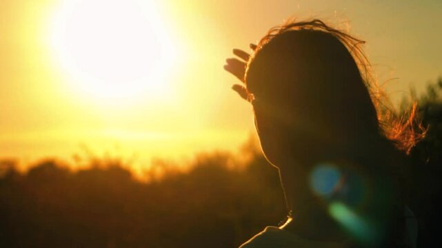 A young beautiful girl looks at the setting sun and covers her face with her hand from the bright rays. 