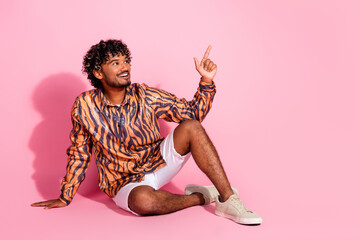 Young man with curly hair sitting on a pink background in a stylish shirt, pointing upward with a...