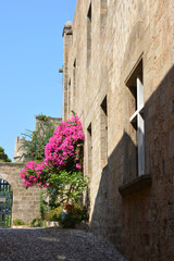A sunny street in Rhodes, Greece, with a historic building and vibrant pink bougainvillea flowers