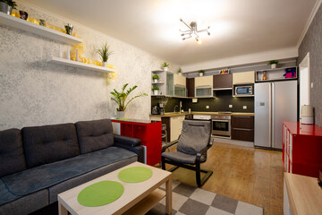 Apartment interior. Living room combined with kitchen. Furniture and kitchen appliances: dark gray sofa, armchair, coffee table, red chests of drawers, chandelier, kitchen set and refrigerator.