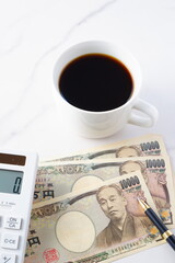 福沢諭吉の肖像の一万円紙幣と電卓、ペン、淹れたてのコーヒーが入ったコーヒーカップ
