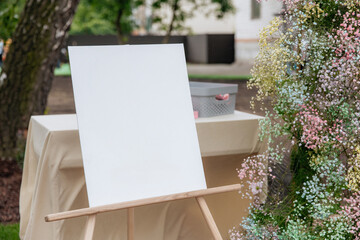 Empty white easel for text and drawing on a background of a garden with flowers. Mockup for artist's creativity