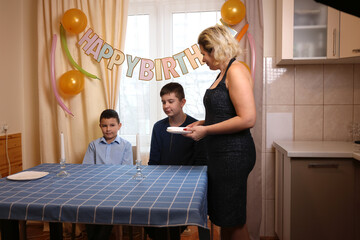 A woman with her son and nephew decorate a room and a festive table for a birthday celebration. Soft lighting.