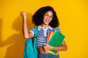 Young cheerful woman celebrating success holding notebooks and a backpack against a vibrant yellow...