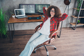 Young woman programmer with curly hair relaxing in her home workspace with computer setup and...