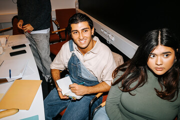 High angle view of smiling young man wearing bib overalls taking notes while sitting near female friend in classroom at