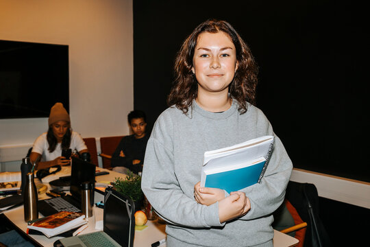 Portrait of smiling young female student holding books while standing near table in classroom at university