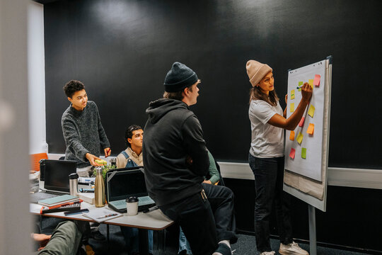 Young female student writing on adhesive note while standing near whiteboard at meeting with friends in university class