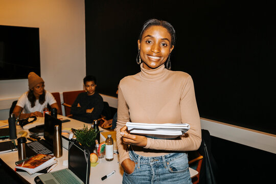 Portrait of happy young female student holding books while standing with hand in pocket near table at college classroom