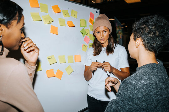 Young woman wearing knit hat standing near whiteboard while talking with male classmate in university