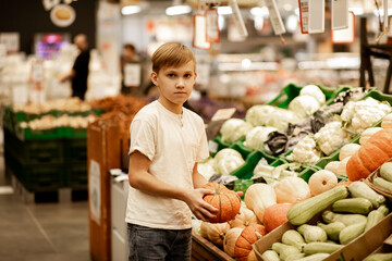 Kid teenager shopping for fresh organic fruits and vegetables in supermarket. Cute boy holding pumpkin in a hands.