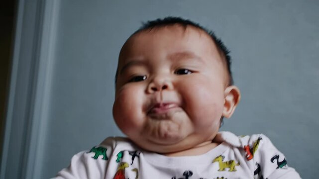 Adorable baby wearing a dinosaur printed onesie puffs out their cheeks and makes a series of playful and funny faces, expressing a range of emotions from happiness to concentration