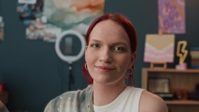 Close-up portrait of positive Caucasian red-haired girl smiling for camera and covering lens with hand in modern room