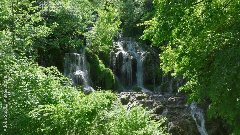 Poster multi-level waterfall flows over mossy rocks surrounded by dense forest. sunlight filters through th