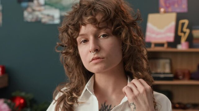 UGC shot of curly-haired gen Z girl with contact lenses, pierced nose and chest tattoo wearing white blouse posing for camera getting ready to start live-streaming at home