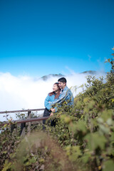 A couple is standing on a wooden bridge, looking out over a lush green forest