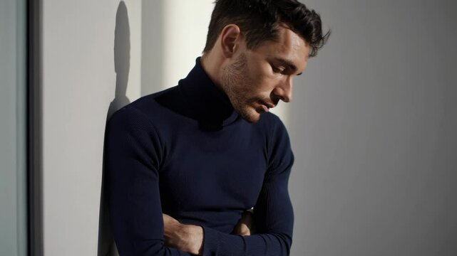 Pensive male model standing with arms crossed, gazing downward in contemplative studio portrait, captured through slow motion cinematography revealing subtle emotional depth