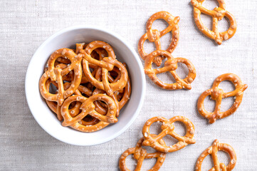 Salted pretzels in a white bowl on linen fabric. Golden toasted small pretzels of traditional shape with intertwined ends to form a pretzel bow, made of wheat flour, used for snacking and dipping.