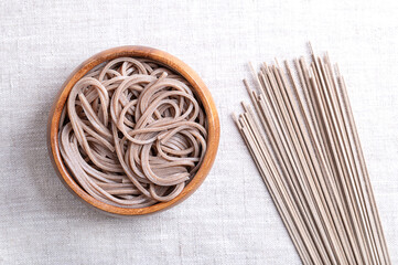 Chilled and boiled soba noodles in a wooden bowl on linen fabric. Thin Japanese noodles, made primarily from buckwheat flour, with wheat flour mixed in. With dried noodles placed on the right side.