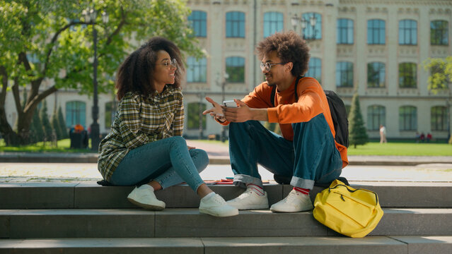 Two happy African American friendly students conversation sitting outdoor multiracial friends African American woman girl female with boy man guy in city talking smiling university college high school