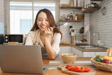 Happy young woman video calling with friends while sitting in kitchen surrounded by fresh...