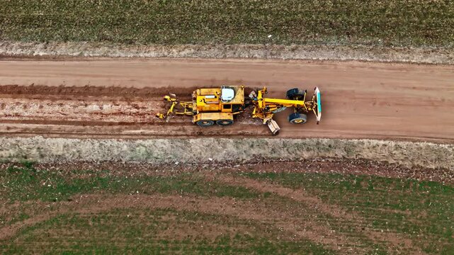 Heavy yellow grader leveling dirt road, aerial top down view