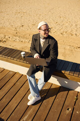 Stylish man with tattoos enjoys a leisurely day at the beach while working on his laptop