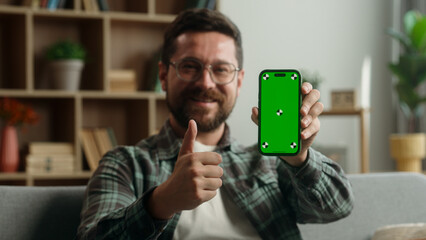 Caucasian man male guy smiling showing green screen smartphone display app at home demonstrate thumb up recommend like good online shopping website chroma key mobile phone bet gambling sport casino