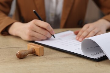 Woman signing licensing agreement document at wooden table indoors, focus on stamp