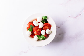 Juicy caprese salad with baby mozzarella cheese, cherry tomatoes and parsley in white ceramic plate on white marble table top view. Healthy diet food concept.