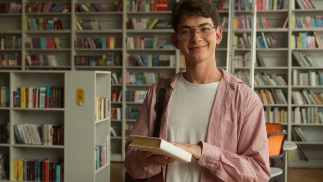 Teenage guy smiling happy looking at camera holding book buying literature prepare for reading in books store library in high school college studying learning Caucasian man male student education