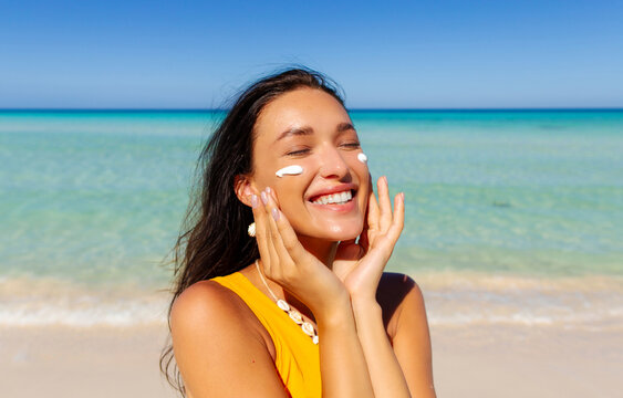 Portrait of beautiful woman applying sun cream on face, standing on sandy beach with turquoise water on background. Skin care, sun protection concept